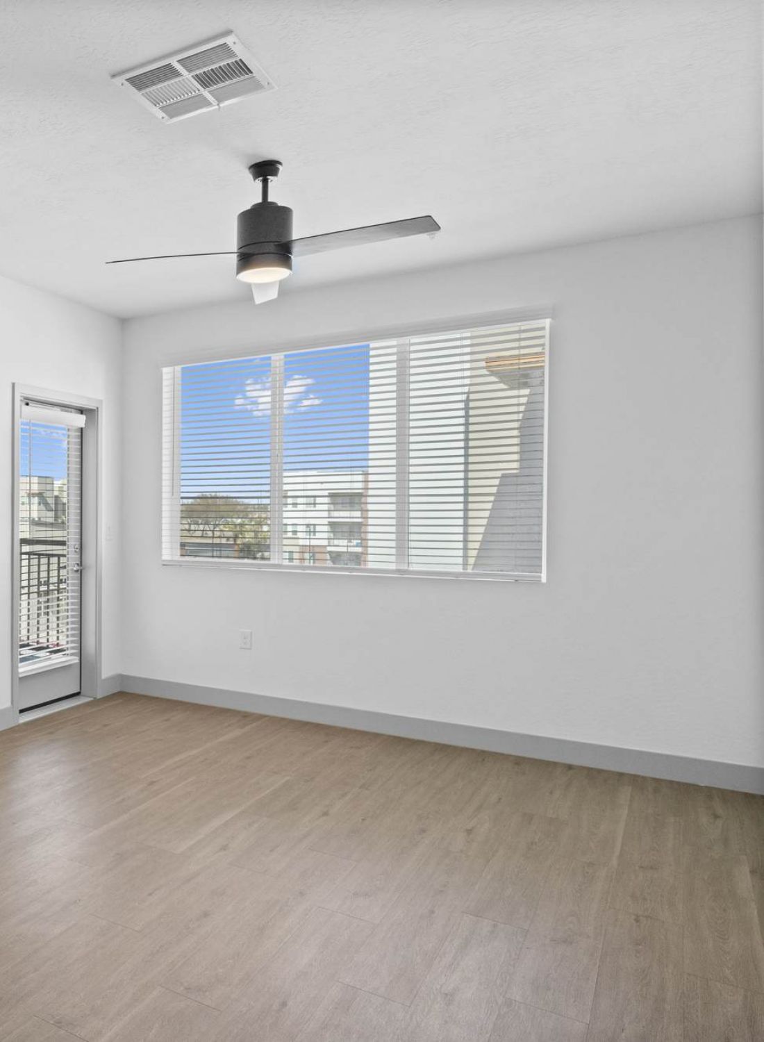 Bright, empty room with wood flooring, ceiling fan, and large window next to a door leading to a balcony.