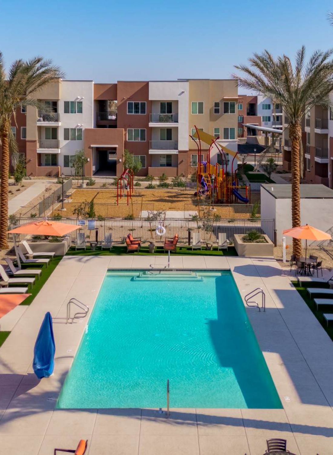 A modern apartment complex with a central swimming pool, lounge chairs, palm trees, and a playground in the background.