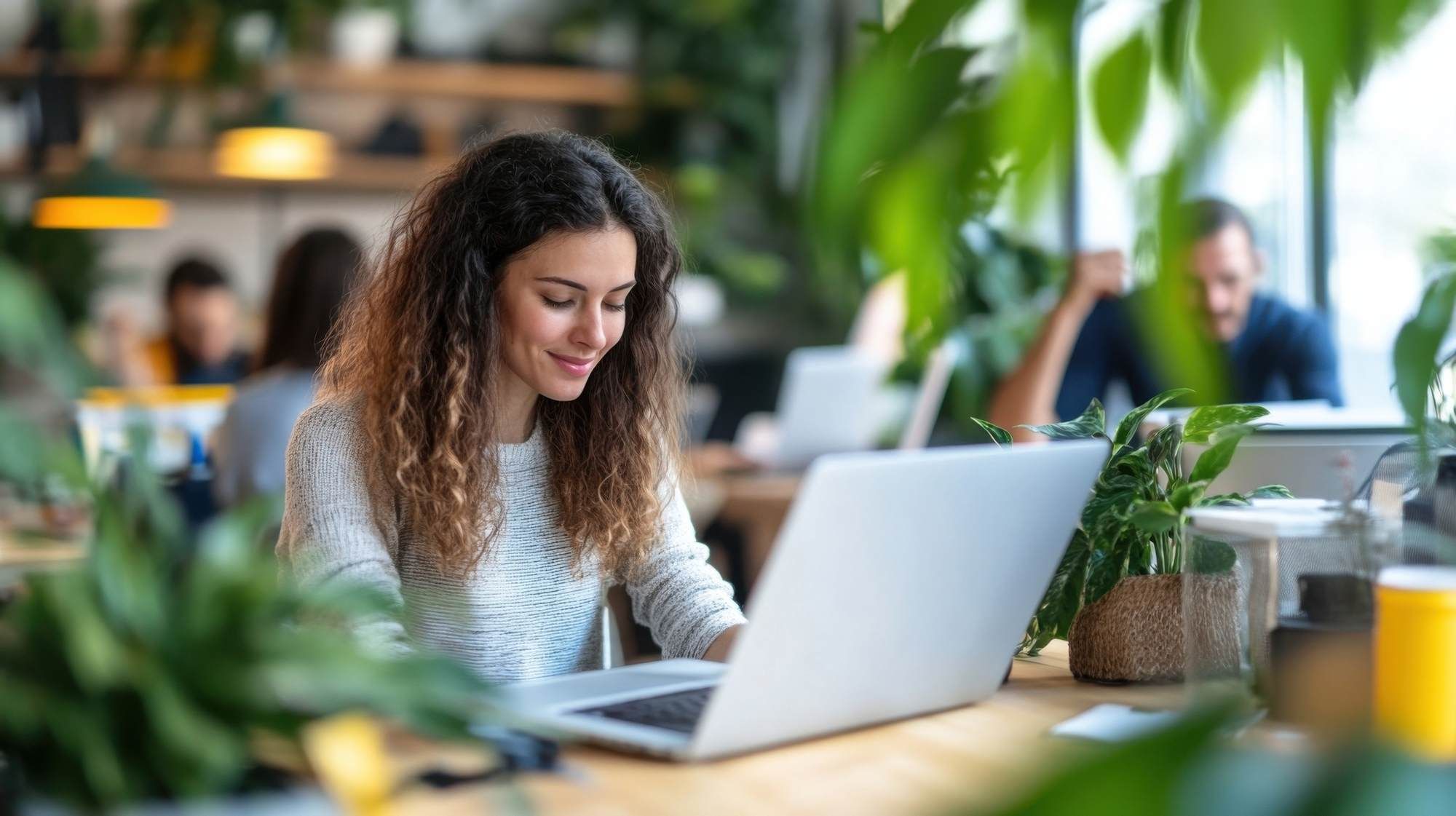 Woman with curly hair working on a laptop at a desk surrounded by plants in a bright, modern workspace.