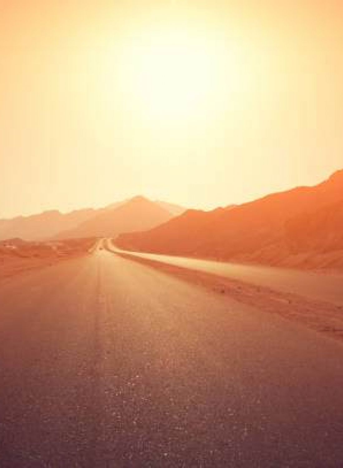 A deserted road in a desert landscape with mountains and a bright, orange-tinted sky at sunset or sunrise.