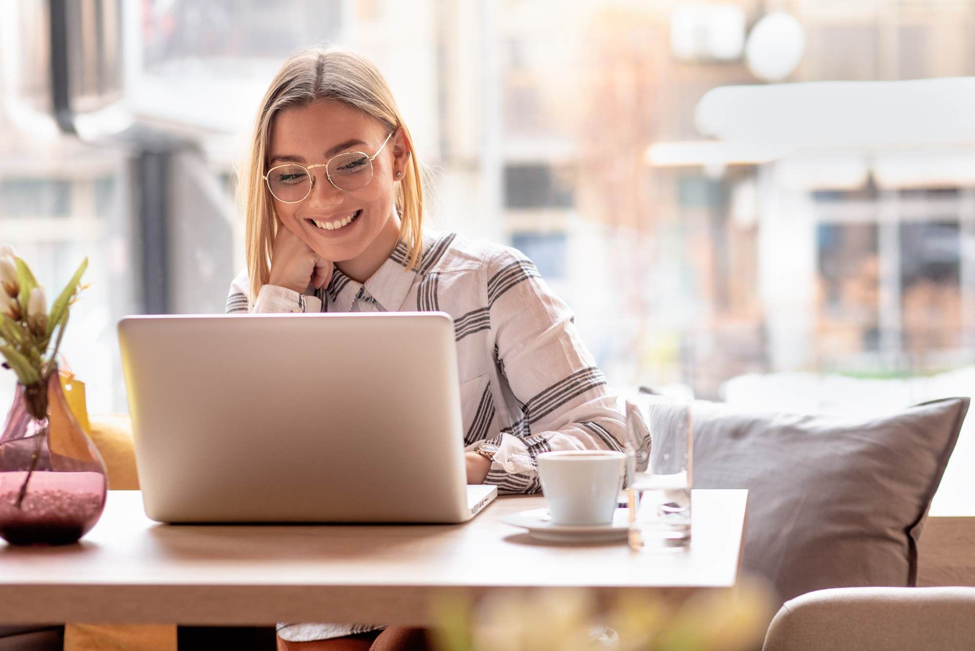 Smiling woman with glasses using a laptop at a cozy café table with a cup of coffee.
