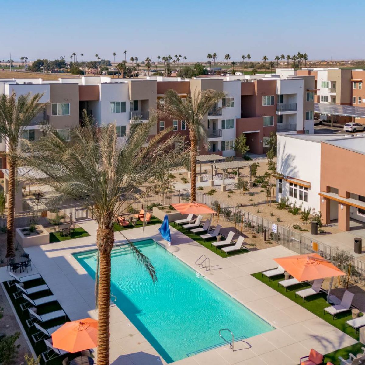 A modern apartment complex with a central pool, palm trees, lounge chairs, and orange umbrellas.