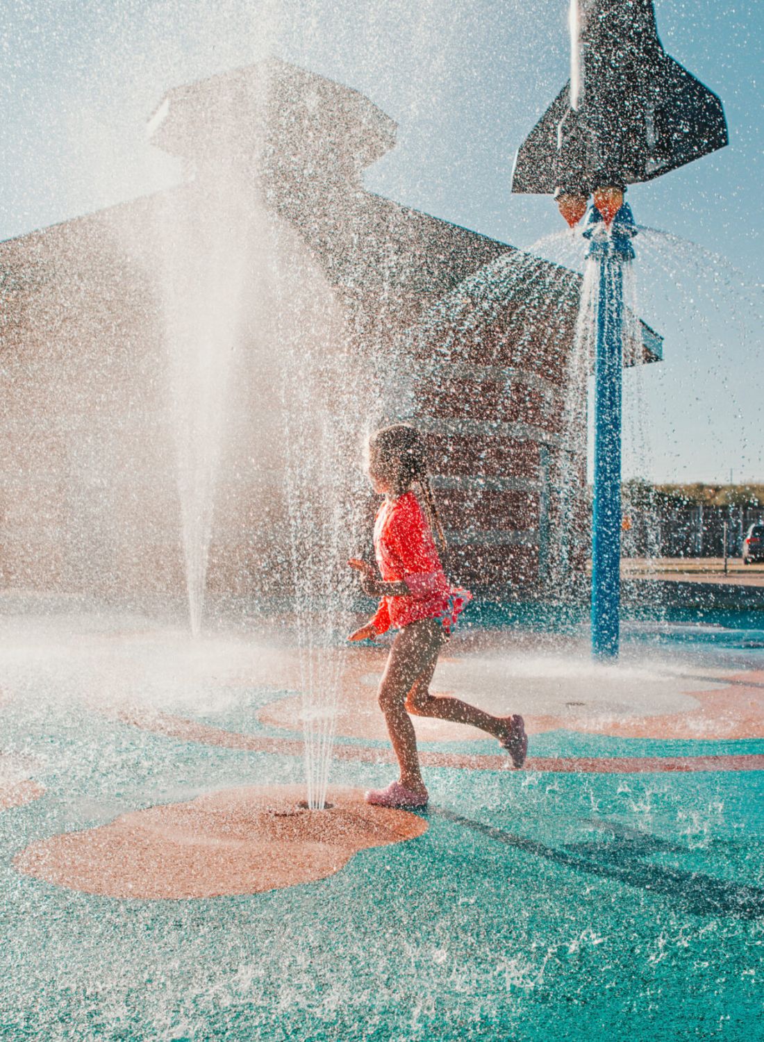 A child runs through water sprays at a splash pad on a sunny day, near a rocket-shaped fountain.
