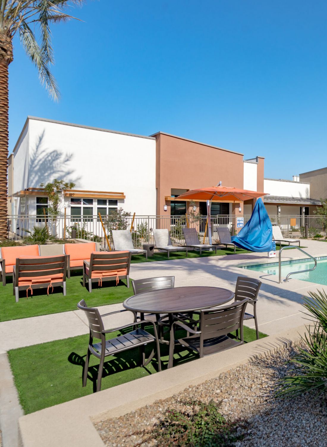 Outdoor pool area with lounge chairs, tables, umbrellas, palm trees, and modern apartment buildings under a blue sky.