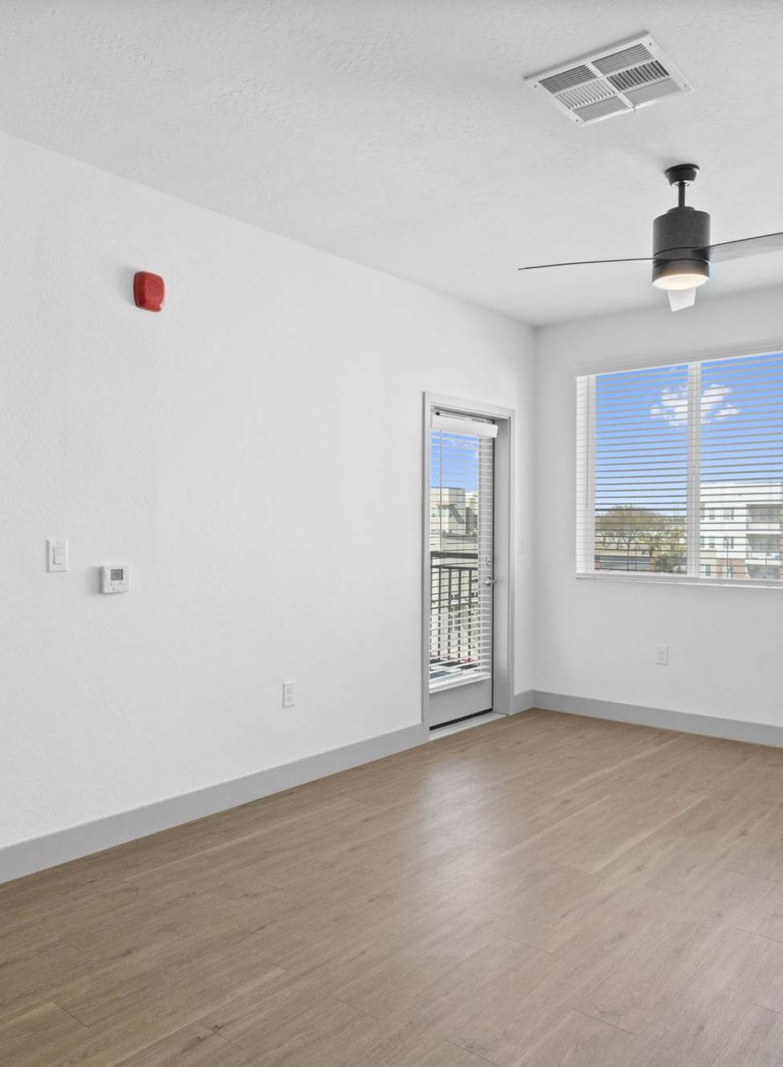 Bright, empty room with wood flooring, ceiling fan, and large window next to a door leading to a balcony.